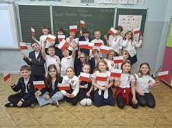 Na zdjęciu widać grupę dzieci z klas 1–3 stojących i siedzących w sali lekcyjnej. Uczniowie są ubrani odświętnie – dominują białe koszule, granatowe i czarne elementy stroju oraz biało-czerwone kotyliony przypięte do ubrań. Każde dziecko trzyma w ręku małą polską flagę, którą dumnie prezentuje. Dzieci pozują razem do zdjęcia, uśmiechają się i wyglądają na radosne oraz zaangażowane w szkolne wydarzenie. W tle znajduje się tablica, na której zapisano temat zajęć związany ze Świętami Majowymi oraz datę. Widać także materiały edukacyjne, m.in. planszę z alfabetem. Całość oddaje uroczystą, ale jednocześnie pogodną atmosferę wspólnego świętowania i nauki o ważnych wydarzeniach historycznych.