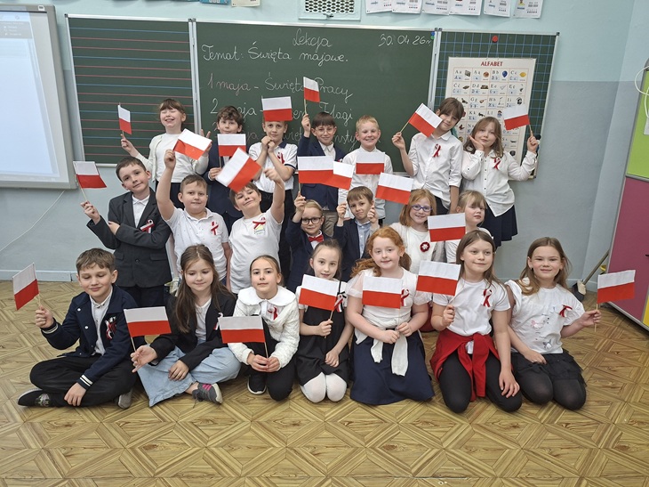 Na zdjęciu widać grupę dzieci z klas 1–3 stojących i siedzących w sali lekcyjnej. Uczniowie są ubrani odświętnie – dominują białe koszule, granatowe i czarne elementy stroju oraz biało-czerwone kotyliony przypięte do ubrań. Każde dziecko trzyma w ręku małą polską flagę, którą dumnie prezentuje. Dzieci pozują razem do zdjęcia, uśmiechają się i wyglądają na radosne oraz zaangażowane w szkolne wydarzenie. W tle znajduje się tablica, na której zapisano temat zajęć związany ze Świętami Majowymi oraz datę. Widać także materiały edukacyjne, m.in. planszę z alfabetem. Całość oddaje uroczystą, ale jednocześnie pogodną atmosferę wspólnego świętowania i nauki o ważnych wydarzeniach historycznych.