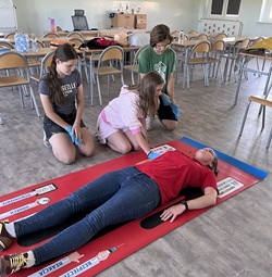 Na zdjęciu uczniowie biorą udział w praktycznych zajęciach z zakresu udzielania pierwszej pomocy. W specjalnie przygotowanej sali ćwiczą postępowanie z osobą poszkodowaną, wykorzystując do tego matę edukacyjną oraz sprzęt szkoleniowy. Trójka uczniów, wyposażona w rękawiczki ochronne, wykonuje kolejne czynności ratunkowe pod okiem nauczyciela odgrywającego rolę osoby poszkodowanej. W tle widoczny jest fantom do nauki resuscytacji krążeniowo-oddechowej, co świadczy o kompleksowym charakterze szkolenia. Zdjęcie pokazuje zaangażowanie uczestników oraz ich odpowiedzialne podejście do nauki umiejętności, które mogą okazać się niezwykle cenne w sytuacjach zagrożenia zdrowia i życia.