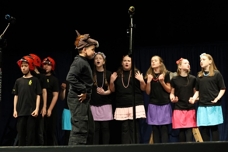 Na zdjęciu widać grupę dzieci występujących na scenie podczas przedstawienia teatralnego. Uczniowie ubrani są w czarne stroje sceniczne, a część dziewczynek ma kolorowe spódniczki i ozdoby we włosach. Po lewej stronie stoją chłopcy w czerwonych hełmach strażackich, co sugeruje, że spektakl dotyczy pracy strażaków lub akcji ratunkowej.  W centralnej części sceny znajduje się chłopiec w kostiumie przypominającym zwierzę lub bajkową postać, który odgrywa główną rolę i zwraca uwagę swoją postawą oraz strojem. Pozostali uczestnicy śpiewają lub recytują swoje kwestie, gestykulując i angażując się w występ.  Na scenie widoczne są mikrofony oraz ciemne tło kurtyny teatralnej, które podkreślają charakter wydarzenia artystycznego. Zdjęcie oddaje atmosferę dziecięcego spektaklu pełnego emocji, zaangażowania i współpracy.