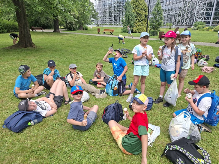 Na zdjęciu widać grupę dzieci odpoczywających na trawie w parku. Większość siedzi lub leży na trawniku, a wiele z nich trzyma lody w wafelkach i je zajada. Dzieci mają na sobie letnie ubrania – koszulki, krótkie spodenki oraz czapki z daszkiem chroniące przed słońcem. Wokół nich leżą plecaki, torby i rower.  W tle znajdują się drzewa, ławki parkowe oraz ścieżka spacerowa. Po prawej stronie widać dużą drewnianą konstrukcję przypominającą fragment kolejki górskiej lub inną dużą atrakcję rekreacyjną. Pogoda wydaje się ciepła i przyjemna, a cała scena sprawia wrażenie wycieczki lub wspólnego wypoczynku grupy dzieci podczas letniego dnia.