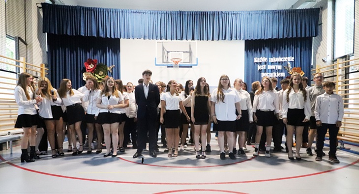 Na zdjęciu uwieczniono uczniów klas ósmych podczas uroczystej akademii z okazji zakończenia szkoły podstawowej. Absolwenci stoją na środku sali gimnastycznej, odświętnie ubrani – w białych koszulach i bluzkach oraz ciemnych spódnicach i spodniach. W centrum grupy znajduje się uczeń w eleganckim garniturze, a pozostali uśmiechają się i z radością uczestniczą w występie.  W tle widoczna jest szkolna scena z granatową kurtyną oraz dekoracja z napisem: „Każde zakończenie jest nowym początkiem”, która doskonale oddaje charakter tego wyjątkowego dnia. Na scenie znajdują się również ozdobne kwiaty i elementy dekoracyjne przygotowane specjalnie na tę okazję.