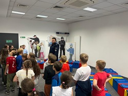 Na zdjęciu widać uczniów klasy 3c podczas spotkania z policjantem w komisariacie. Dzieci słuchają informacji o bezpieczeństwie i pracy funkcjonariuszy. W sali znajdują się materiały edukacyjne związane z pracą policji.