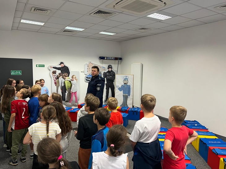 Na zdjęciu widać uczniów klasy 3c podczas spotkania z policjantem w komisariacie. Dzieci słuchają informacji o bezpieczeństwie i pracy funkcjonariuszy. W sali znajdują się materiały edukacyjne związane z pracą policji.