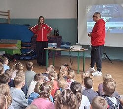 Na zdjęciu widać spotkanie uczniów z przedstawicielami świata sportu. W sali gimnastycznej przed zgromadzonymi dziećmi stoi młoda zawodniczka w stroju sportowym oraz mężczyzna prowadzący spotkanie. W tle znajduje się ekran z prezentacją multimedialną, a obok przygotowany jest sprzęt sportowy.  Uczniowie z zainteresowaniem obserwują rozmowę i prezentację. Zdjęcie pokazuje edukacyjne i inspirujące spotkanie, podczas którego dzieci miały okazję poznać bliżej sport oraz życie młodej zawodniczki.
