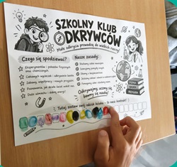 Na zdjęciu widać kartę informacyjną Szkolnego Klubu Odkrywców. Karta znajduje się na ławce, a dziecko wskazuje palcem jeden z jej elementów.  Na plakacie przedstawiono kolorowe, dziecięce ilustracje związane z nauką i odkrywaniem świata, m.in. lupę, żarówkę, probówki oraz postacie młodych odkrywców. Znajdują się na nim również informacje dotyczące działalności Klubu, między innymi tego, czego uczniowie będą poszukiwać i jakie zasady będą obowiązywać podczas zajęć.  Zdjęcie pokazuje ciekawą i angażującą formę zaprezentowania dzieciom zajęć przyrodniczych, zachęcającą je do eksperymentowania, zadawania pytań i odkrywania świata.